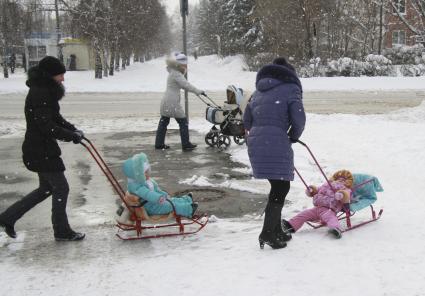 Три женщины гуляют с детьми по городу. Двое детей едут на санках, одного везут в детской коляске.