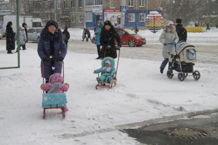 Три женщины гуляют с детьми по городу. Двое детей едут на санках, одного везут в детской коляске.