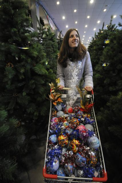 Магазин по продаже новогодних елок. На снимке: Девушка с тележкой полной елочных игрушек выбирает себе искусственную новогоднюю елку.