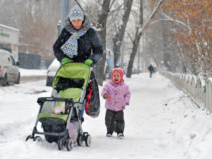 Женщина с детской коляской идет по заснеженной улице, рядом бежит ребенок в слезах.