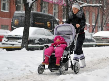 Женщина с ребенком в детской коляске идет по заснеженной улице.