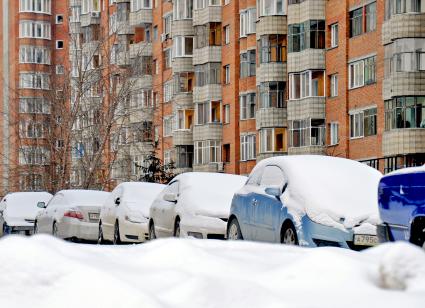 Засыпанные снегом автомобили рядом с жилым домом.