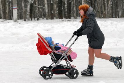 Девушка на коньках катается на катке везя впереди себя детскую коляску.