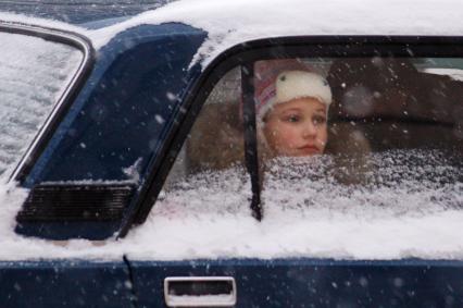 Девочка смотрит из окна засыпанного снегом автомобиля.