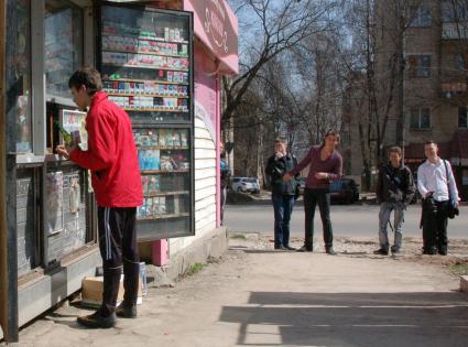 Подросток покупает пиво в уличной палатке. Друзья стоят в стороне, наблюдают за процессом.