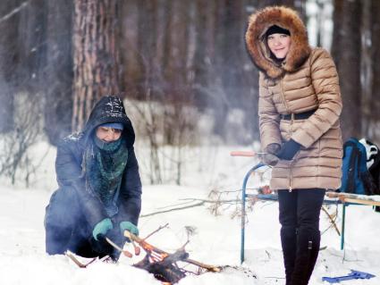 Две девушки готовят на костре сосиски в зимнем лесу.