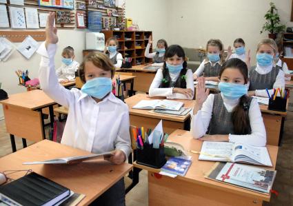 Школьники в медицинских повязках поднимают руки на уроке.