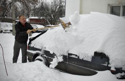 Мужчина отчищает автомобиль от снега с помощью деревянной лопаты.