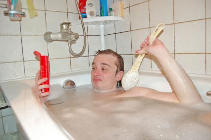 Парень, лежащий в ванне с пеной, держит флакон с моющим средством и щетку.