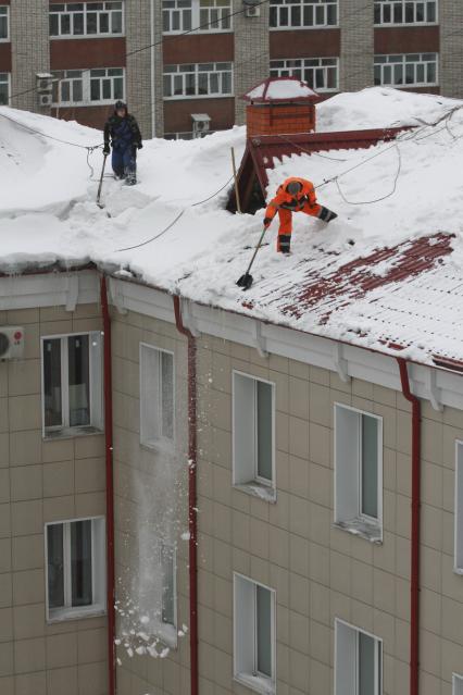 Работник службы ЖКХ в ярком оранжевом комбинезоне чистит лопатой крышу от снега прикрепив себя страховочным тросом.