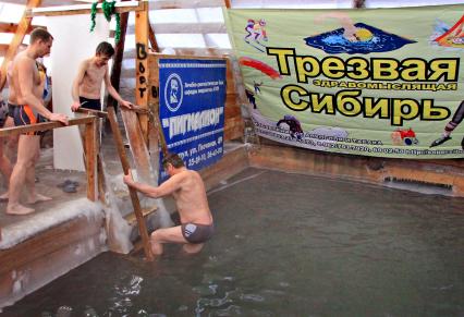 Мужчины окунаются зимой в прорубь во время празднования христианского праздника крещение, на фоне плаката с надписью: `Трезвая здравомыслящая Сибирь`.