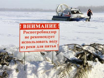 На льду водоема стоит судно на воздушной подушке МЧС `Марс 700` на фоне таблички: `Внимание! Роспотребнадзор не рекомендует использовать воду из реки для питья`.