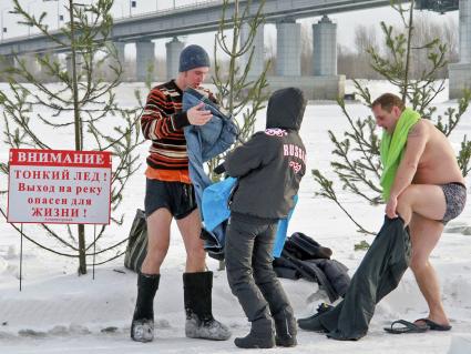 Люди зимой на берегу замерзшей реки одеваются после окунания в прорубь во время праздника крещения, рядом стоит табличка с надписью: `Внимание! Тонкий лед! Выход на реку опасен для жизни`.