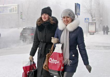 Две девушки улыбаясь идут под руку по заснеженной улице Барнаула.