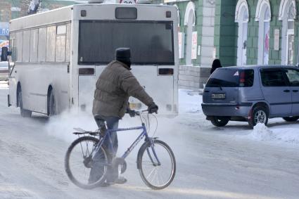 Велосипедист зимой стоит на дороге, рядом стоит автомобиль и проезжает автобус.