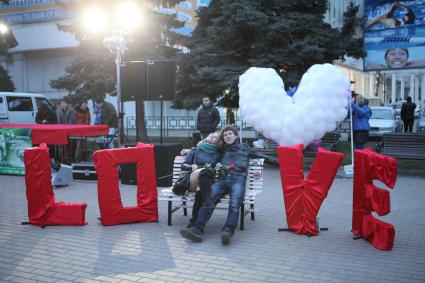 Юноша и девушка с розой в руке сидят на скамейке среди букв: `Love`.