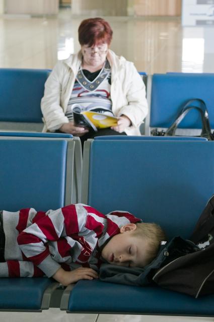 В ожидании рейса ребенок спит на кресле в аэропорту, женщина читает журнал.