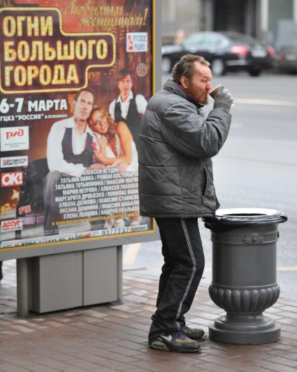 Бомж пьет кофе на улице рядом с урной.