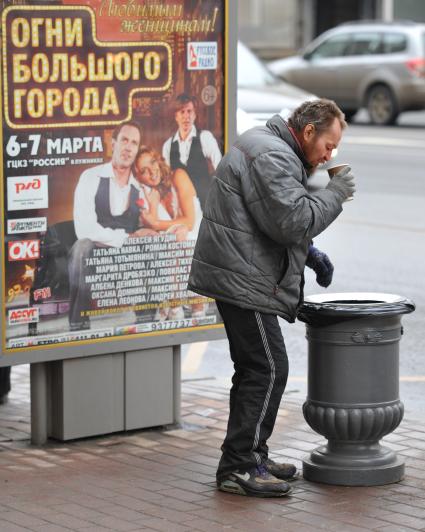 Бомж пьет кофе на улице рядом с урной.