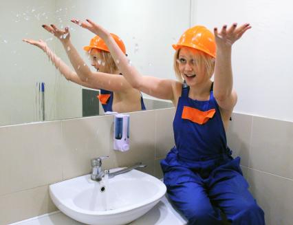 Девушка в спецодежде и каске занимается ремонтом водопроводного крана в ванной комнате.