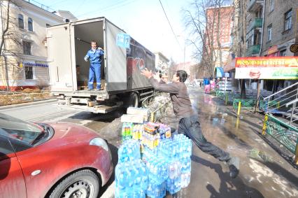 Рабочие выгружают из машины упаковки с водой и напитками. Магазин `Куриный дом` (справа).