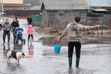 Мужчина с ведром воды идет по затопленной улице, на него смотрят жители села.