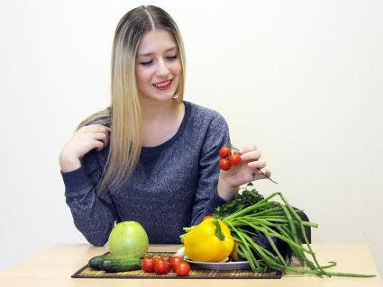 Девушка сидит за столом с овощами и фруктами и держит в руках томат.