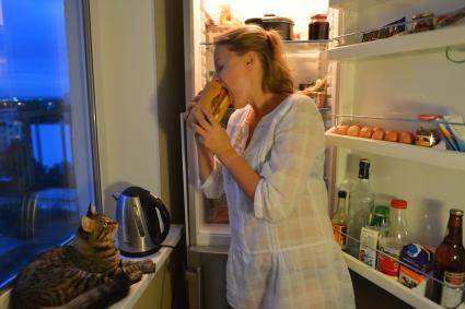 Девушка ест украдкой бутерброд у холодильника.