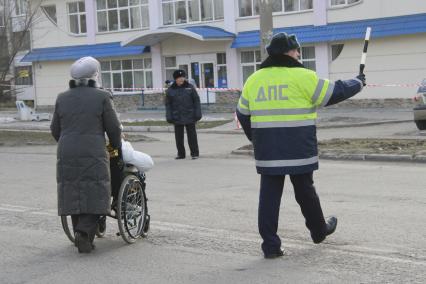 Сотрудник ГИБДД помогает перейти дорогу женщине везущей инвалида в коляске.