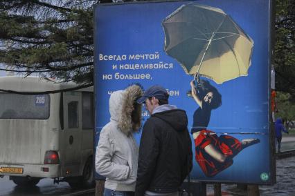 Юноша и девушка разговаривают на улице напротив рекламы с надписью: `Всегда мечтай и нацеливайся на большее`.