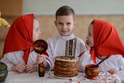 Масленица. На снимке: девочки-близняшки и мальчик в русских народных костюмах сидят за столом с блинами.