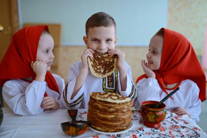 Масленица. На снимке: девочки-близняшки и мальчик в русских народных костюмах сидят за столом с блинами.