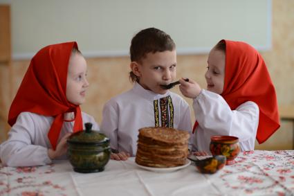 Масленица. На снимке: девочки-близняшки и мальчик в русских народных костюмах сидят за столом с блинами.