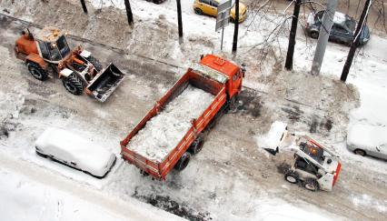 Трактор и минипогрузчик загружают снег в грузовкик `Камаз`.