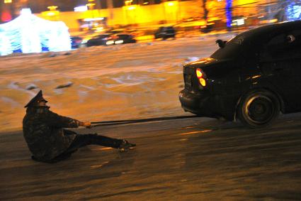 молодой человек в форме полицейского по льду катается на веревке, привязанной к заднему бамперу автомобиля.
