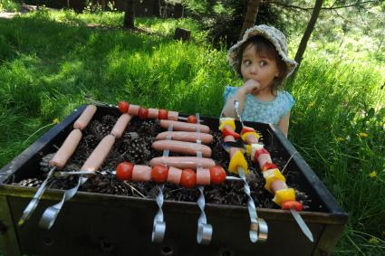 Маленькая девочка у мангала с шашлыком из сосисок.