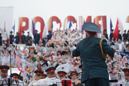 Дирижер под дождем руководит оркестром и сводным зором во время выступления на праздновании дня России