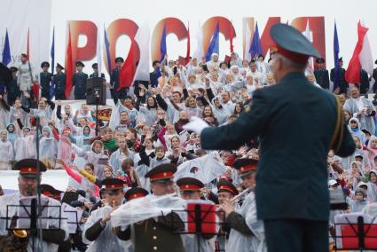 Дирижер под дождем руководит оркестром и сводным зором во время выступления на праздновании дня России