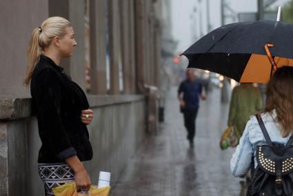 Девушка стоит у стены во время дождя когда мимо проходят люди с зонтами