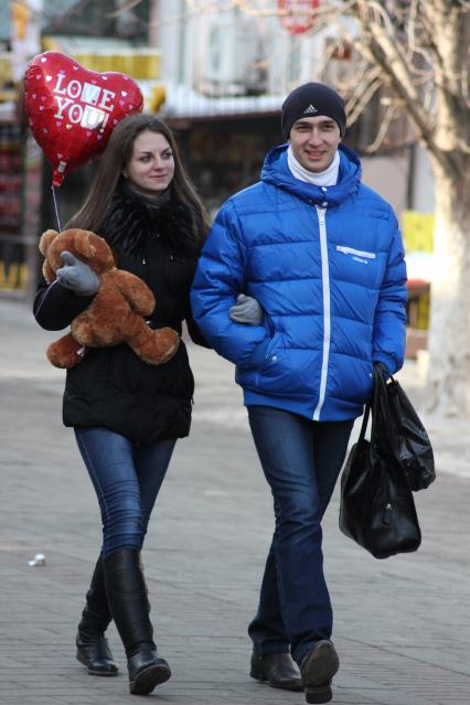 Мужчина и женщина идут подруку. У женщины в руках плюшевый медведь и надувной шар в виде сердца.