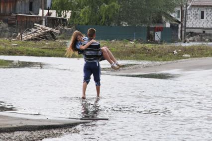 Парень несет на руках девушку через затопленный участок дороги.