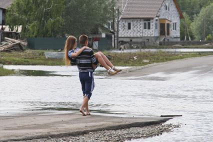 Парень несет на руках девушку через затопленный участок дороги.