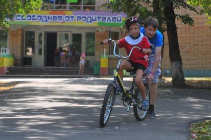 Сеть лагерей  КИД-клуб KIDS. Подмосковный лагерь \'Контакт\'. На снимке: подросток учит мальчика кататься на велосипеде.