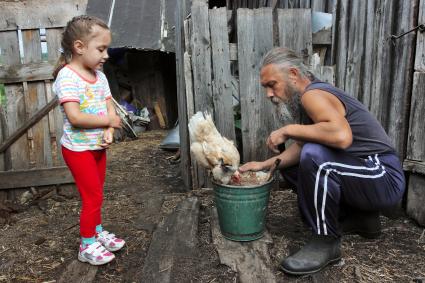 Мужчина кормит курицу, за ним наблюдает ребенок.