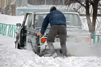 Мужчина толкает увязший в снегу автомобиль.