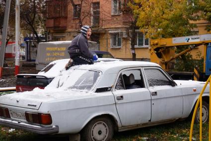 мальчик играет в снежки сидя на крыше старой машины Волга, на детской площадке во дворе жилого дома