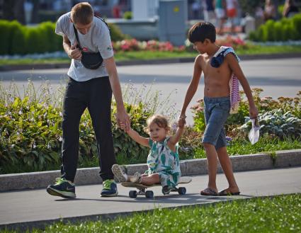 Мужчина с мальчиком везут девочку на скейтборде.