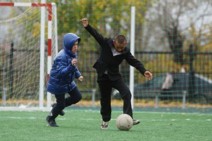 Мальчик в пиджаке, брюках и кросовках играет в футбол на школьном стадионе