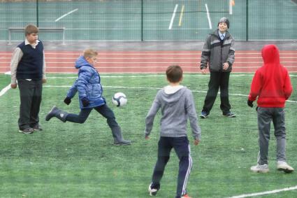 Мальчик в резиновых сапогах играет в футбол на школьном стадионе