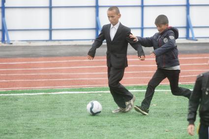 Мальчик в пиджаке, брюках и кросовках играет в футбол на школьном стадионе
